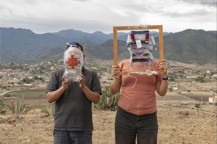 Two artists hold up tapestry weavings in front of their faces against a backdrop of a Californian town in the desert 