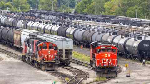 Railway cars are densely packed at the CN Rail freight depot in Hamilton, Ontario, Canada. Two red CN locomotives are visible in the foreground, surrounded by numerous black tanker cars and other freight cars. Trees and greenery are visible in the background