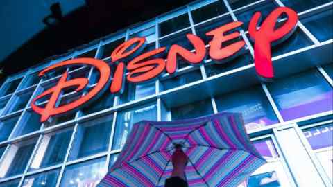 A large red neon sign reading "Disney" is prominently displayed on the exterior of a building. Below the sign, a person holds a multicolored striped umbrellaSquare, New York.