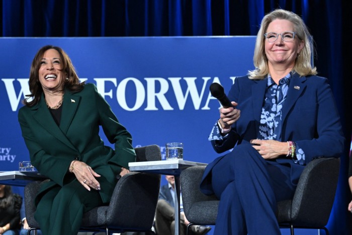 Kamala Harris and Liz Cheney sit on stage during a town hall discussion. Harris is wearing a green suit and laughing, while Cheney, in a blue suit, holds a microphone and smiles